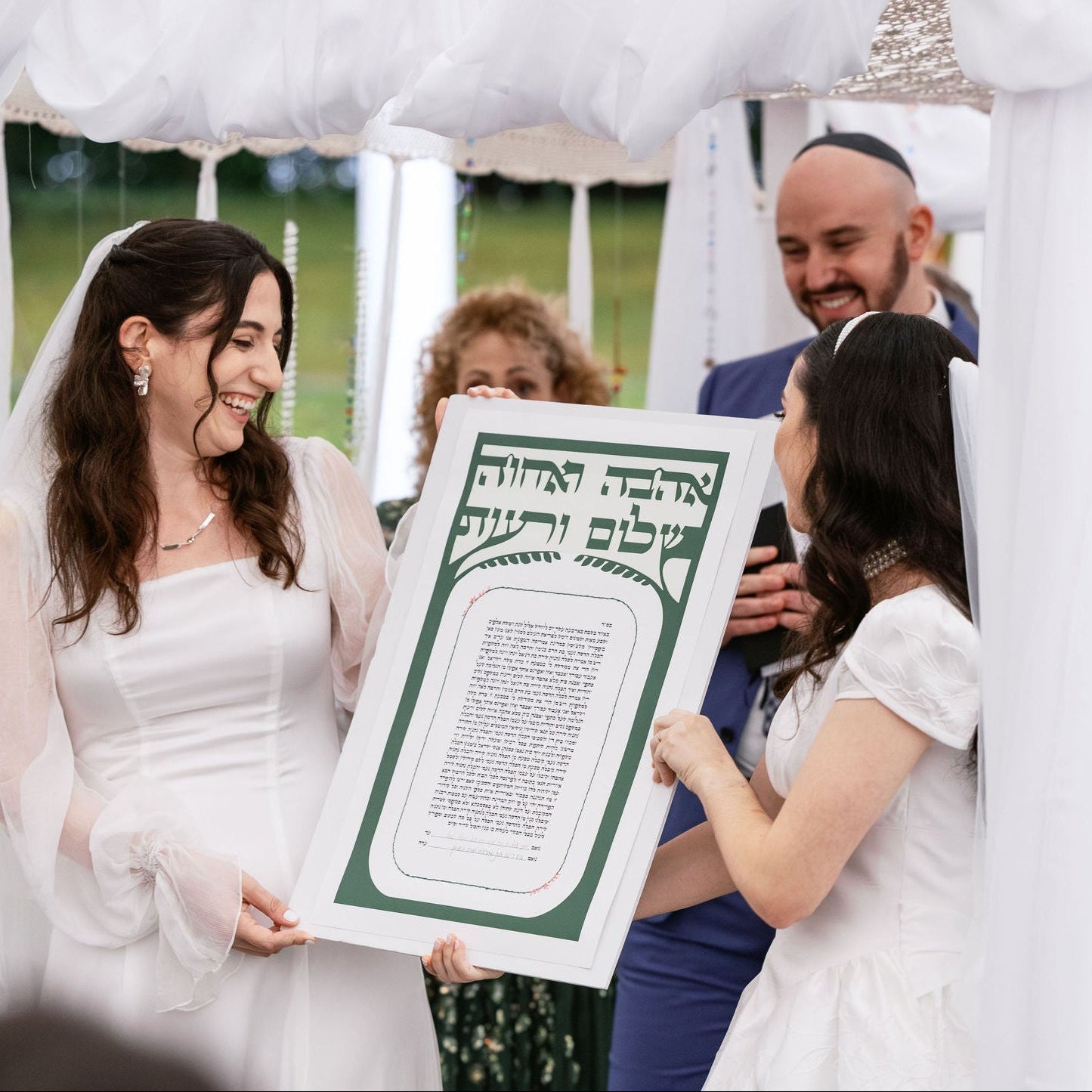 Two brides holding a large framed Hebrew and English marriage contract (Ketubah) under a white fabric chuppah, with a rabbi and guests in the background.