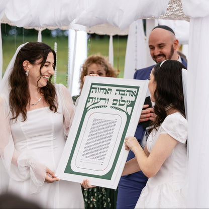 Two brides holding a large framed Hebrew and English marriage contract (Ketubah) under a white fabric chuppah, with a rabbi and guests in the background.