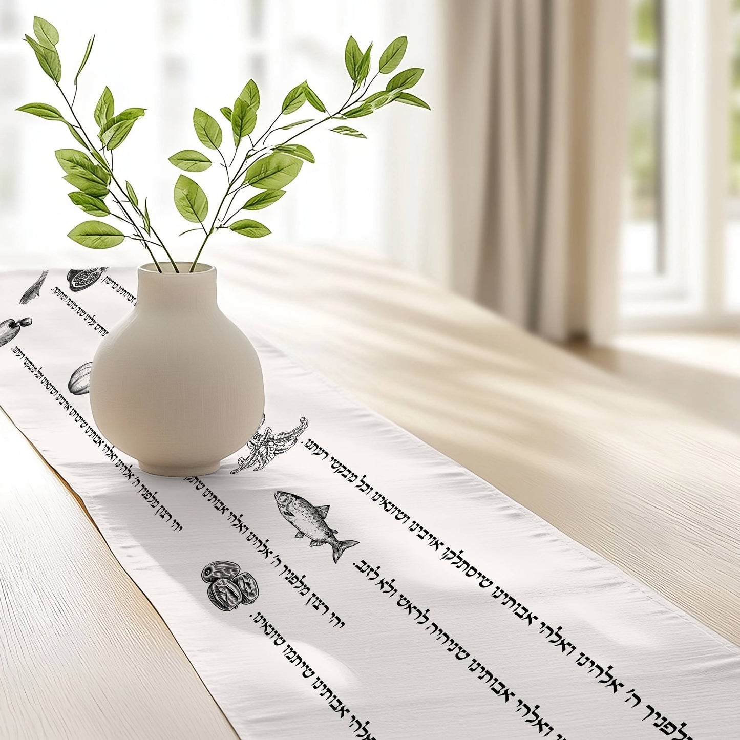 A white table runner with black Hebrew text and decorative fish illustrations, styled on a light wooden table with a small white vase holding green leaves, in a bright, softly blurred room.
