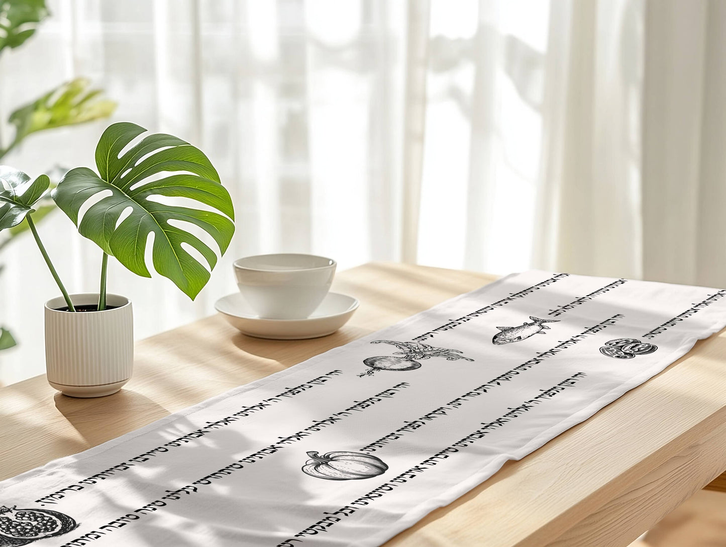 A white table runner with hebrew prayer illustrations laid out on a light wood table in a bright, sunlit room with a potted plant and a white cup on a saucer nearby.