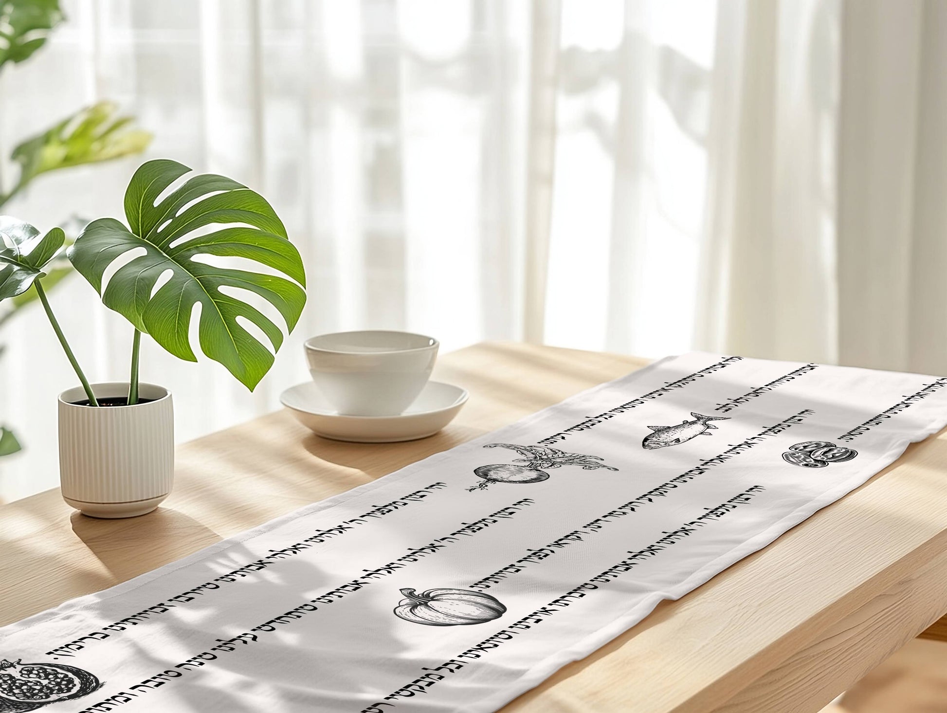 A white table runner with hebrew prayer illustrations laid out on a light wood table in a bright, sunlit room with a potted plant and a white cup on a saucer nearby.