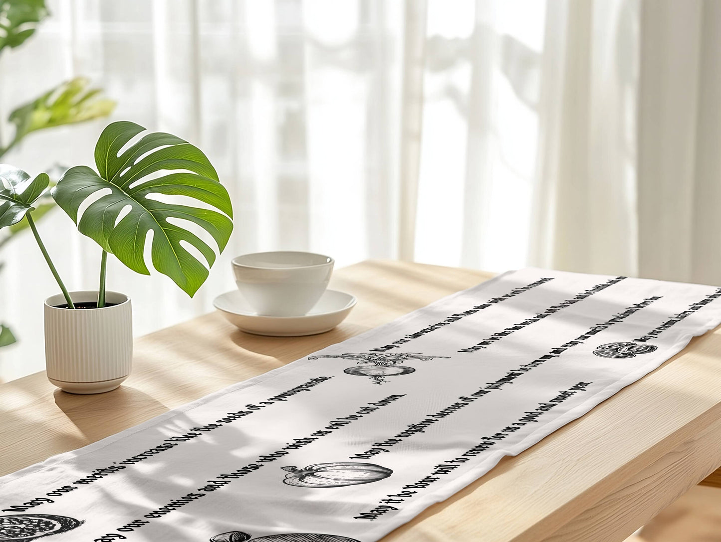 A white table runner with black printed script and decorative motifs laid across a light wood table in a bright, softly lit room. A potted green plant and a white cup and saucer sit on the table, with sheer curtains in the background.