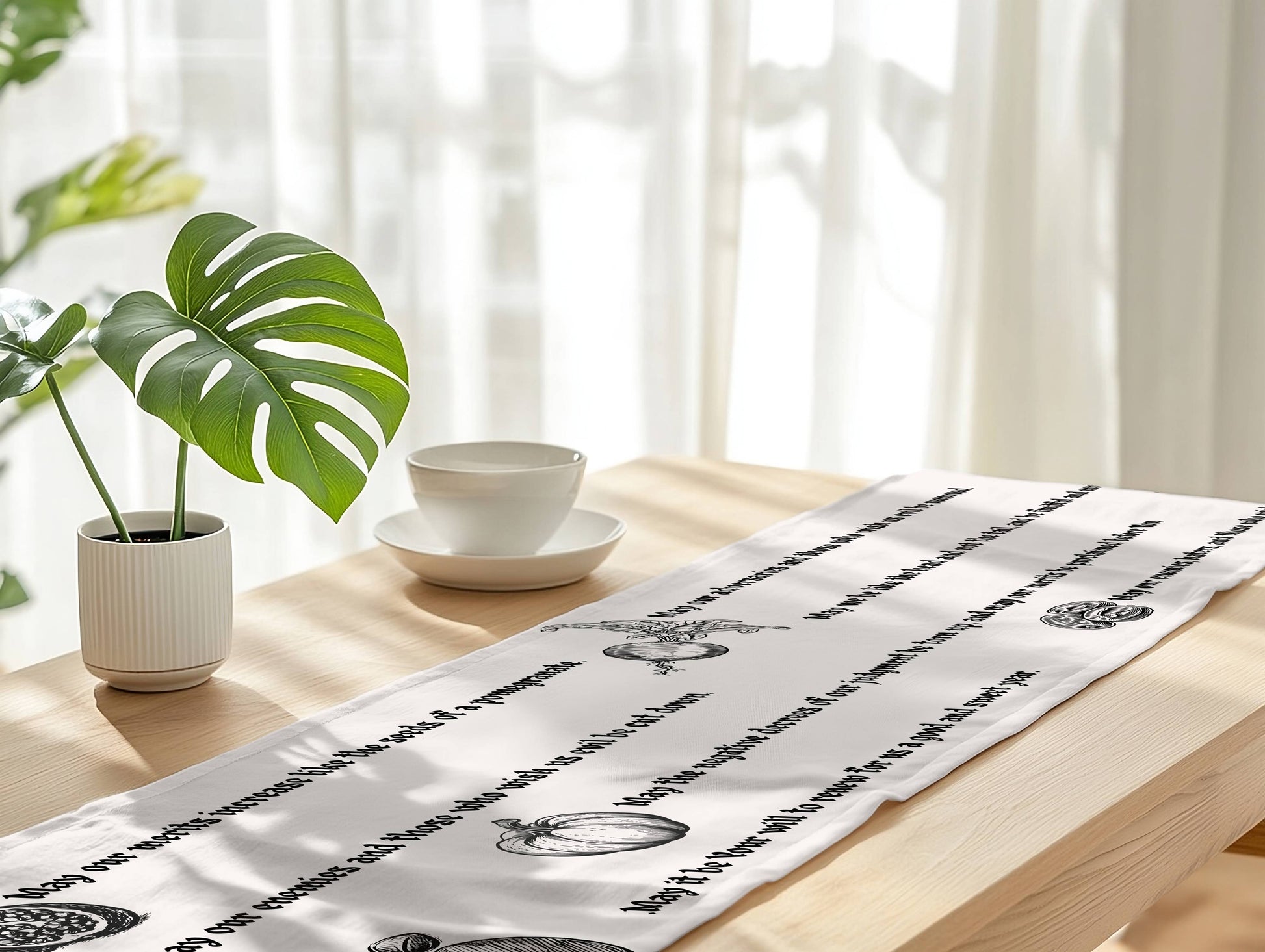 A white table runner with black printed script and decorative motifs laid across a light wood table in a bright, softly lit room. A potted green plant and a white cup and saucer sit on the table, with sheer curtains in the background.