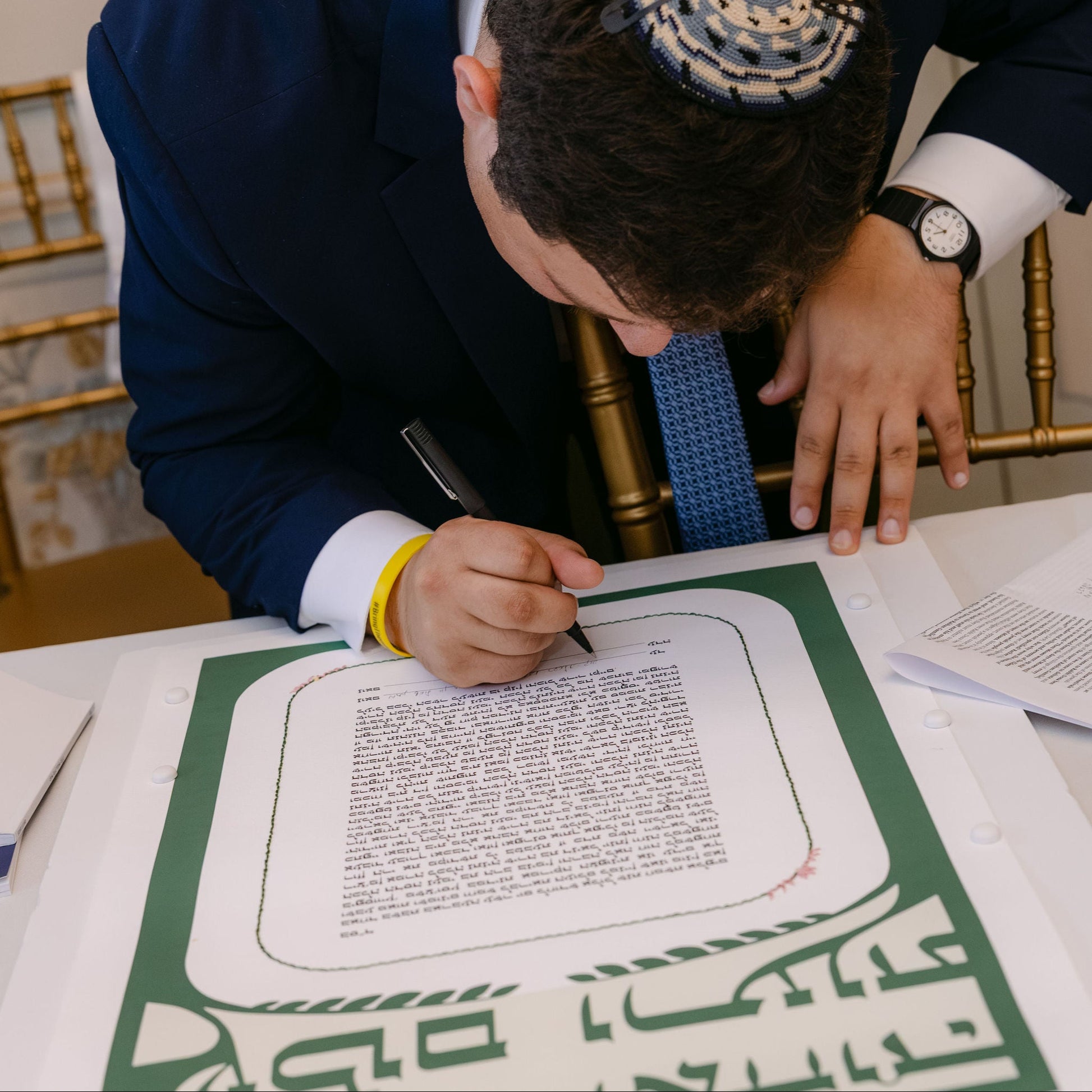 A man in a navy blue suit and patterned kippah leans over a table, writing on a document with a black pen. The document has a green border with Hebrew text.